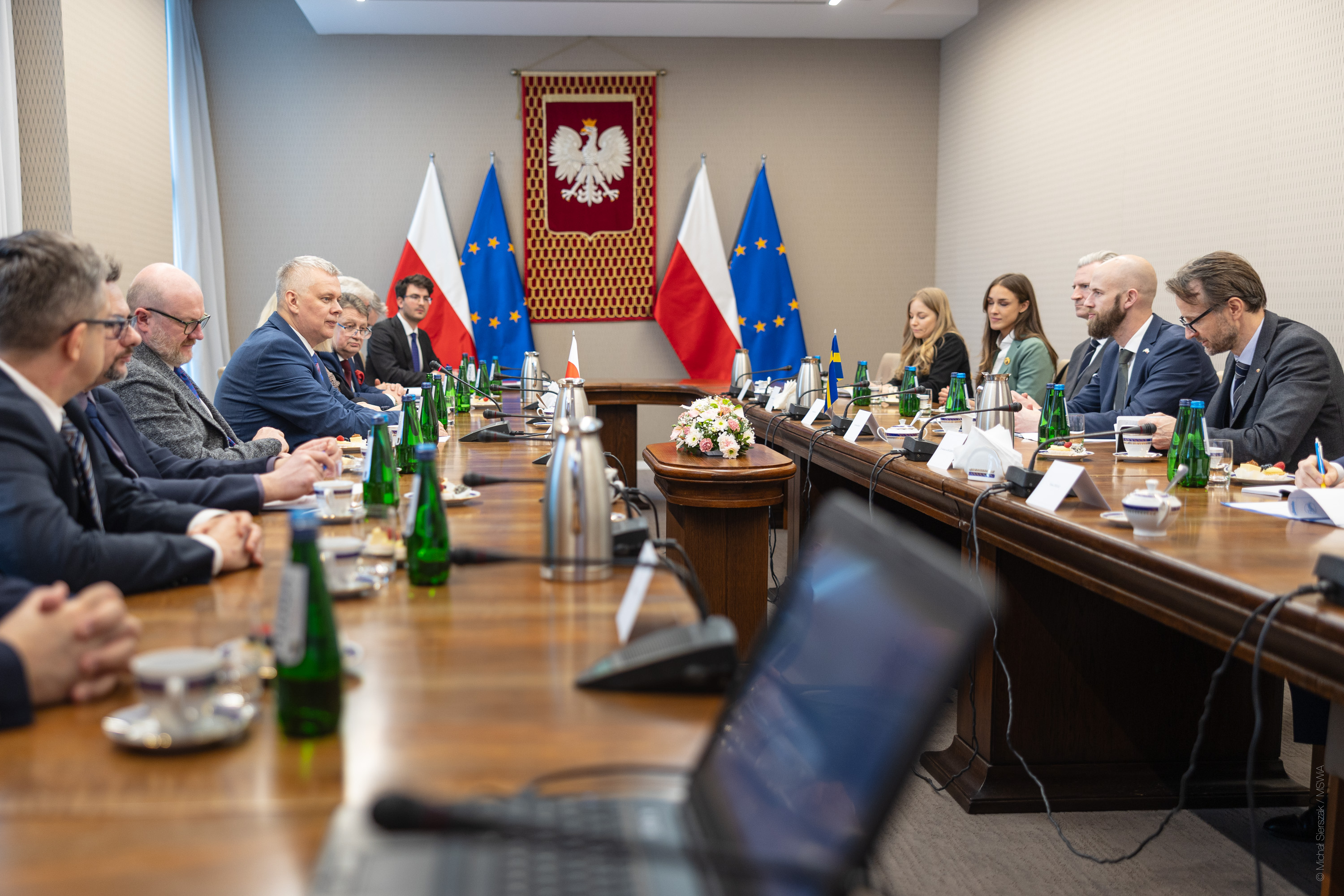 Spotkanie urzędników przy długim stole. W tle godło Polski oraz flagi Polski i UE